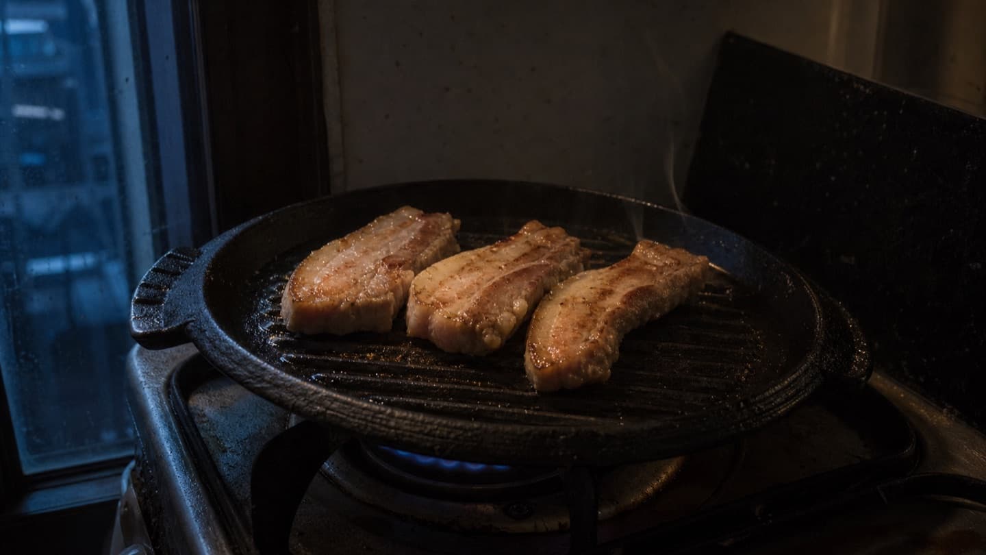 저조도 원룸 주방에서 팬 위 삼겹살이 구워지는 장면. 창문으로 들어온 빛에 옅은 연기가 보인다.